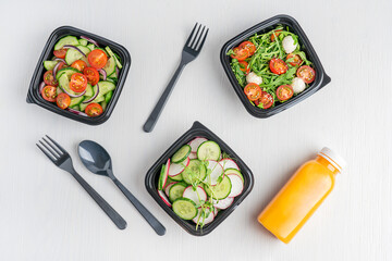 Variety of vegan vegetable salads made of cucumber slices, arugula and tomatoes served in black disposable lunch containers or boxes with orange juice bottle and tableware on white wooden background
