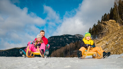 Father with kids on sled riding down snow with beautiful mountain and blue sky behind.