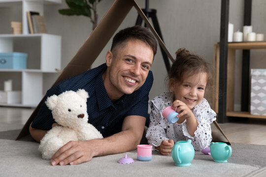 Portrait of happy affectionate young father and small 6s adorable child daughter lying on floor carpet under cardboard roof, imagining playing tea ceremony with favorite toys, family relations concept
