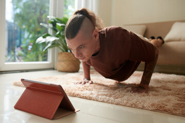 Determined young man keeping feet on sofa when doing push-ups at home and watching tutorial on...