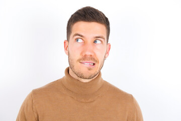 Amazed young caucasian man wearing grey turtleneck over white background bitting lip and looking tricky to empty space.