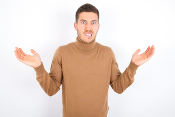 Fototapeta premium Clueless young caucasian man wearing grey turtleneck over white background shrugs shoulders with hesitation, faces doubtful situation, spreads palms, Hard decision