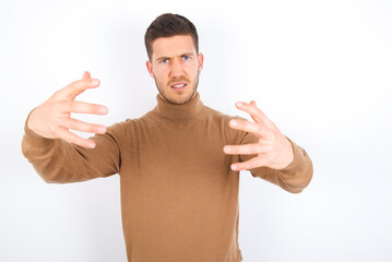 young caucasian man wearing grey turtleneck over white background Shouting frustrated with rage, hands trying to strangle, yelling mad.