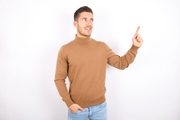 Smiling young caucasian man wearing grey turtleneck over white background indicating finger empty space showing best low prices