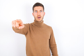 young caucasian man wearing grey turtleneck over white background Pointing with finger surprised ahead, open mouth amazed expression, something on the front.