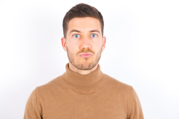 Shot of pleasant looking young caucasian man wearing grey turtleneck over white background , pouts lips, looks at camera, Human facial expressions