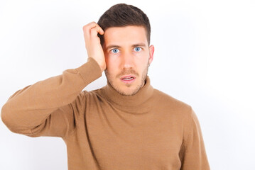 Embarrassed young caucasian man wearing grey turtleneck over white background with shocked expression, expresses great amazement, Puzzled model poses indoor