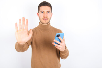 young caucasian man wearing grey turtleneck over white background using and texting with smartphone with open hand doing stop sign with serious and confident expression, defense gesture