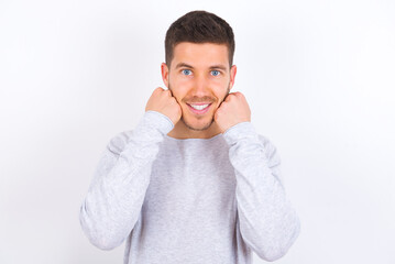 Happy young caucasian man wearing grey sweater over white background keeps fists on cheeks smiles broadly and has positive expression being in good mood