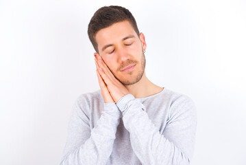 young caucasian man wearing grey sweater over white background leans on pressed palms closes eyes and has pleasant smile dreams about something