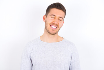 Fototapeta premium Positive young caucasian man wearing grey sweater over white background with overjoyed expression closes eyes and laughs shows white perfect teeth