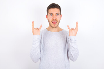 young caucasian man wearing grey sweater over white background makes rock n roll sign looks self...