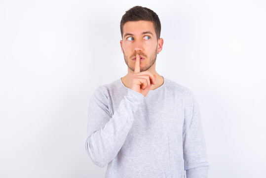 young caucasian man wearing grey sweater over white background silence gesture keeps index finger to lips makes hush sign. Asks not to share secret
