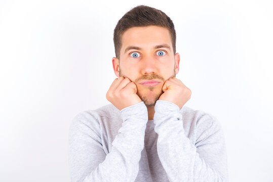 young caucasian man wearing grey sweater over white background with surprised expression keeps hands under chin keeps lips folded makes funny grimace