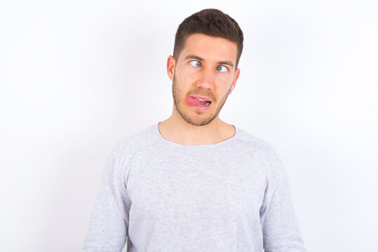 Funny young caucasian man wearing grey sweater over white background makes grimace and crosses eyes plays fool has fun alone sticks out tongue.