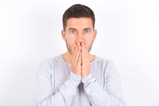 young caucasian man wearing grey sweater over white background keeps hands on mouth, looks with eyes full of disbelief, being puzzled with amount of work