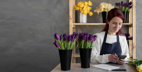 a young female florist in a blue apron checks orders on a mobile phone while sitting at a table with fresh flowers in glass vases in a store. The concept of a small flower delivery business. banner.