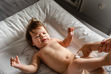 Small caucasian baby girl or boy lying on the bed in bright room while hands of her mother changing diapers and clothes copy space parenting childhood concept cleaning skin wet wipe copy space - obrazy, fototapety, plakaty