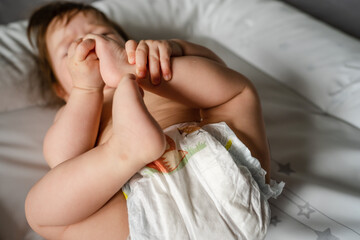 Top view on small caucasian baby lying on her back naked holding legs in diapers on the bed at home with copy space childhood and growing up concept