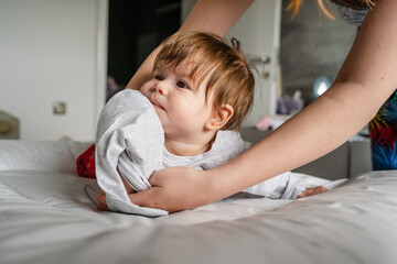 Caucasian baby lying on the bed at home in the bright room on the belly down while her mother is standing behind looking for clothes to change real people childhood and family concept