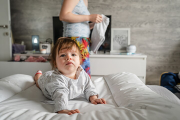 Caucasian baby lying on the bed at home in the bright room on the belly down while her mother is standing behind looking for clothes to change real people childhood and family concept
