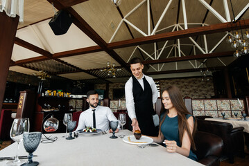 A young waiter in a stylish apron serves a table with a beautiful couple in a refined restaurant. Exquisite delicacies of haute cuisine close-up.