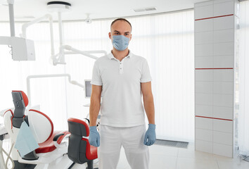 Naklejka na ściany i meble A young dentist in a mask stands near a red dental chair and smiles in modern white dentistry. Modern dentistry and prosthetics.
