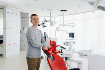 A young man stands near a red dental chair and smiles in modern white dentistry. Treatment and prevention of caries from youth. Modern dentistry and prosthetics.