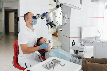 A young dentist examines and treats the teeth of a young guy in modern white dentistry using a...