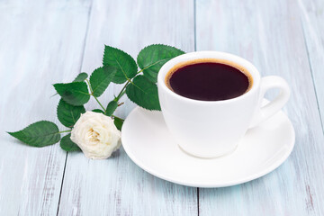 Dark coffee in white cup, beautiful rose on background, light blue wooden table, horizontal