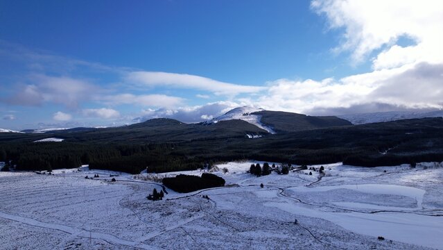 Aerial image of a road through bleak snow-covered moorland. With a dark conifer plantation and a blue sky with white clouds.