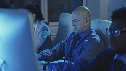 Chest-up of mature intelligent Caucasian man sitting at desk in dark modern office, drinking coffee from paper cup, using computer at night, blurred colleagues working on sides - Powered by Adobe