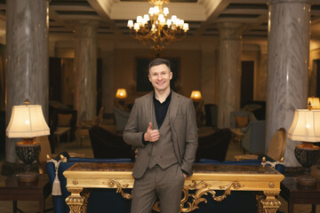 Businessman man in a classic stands in the luxurious interior of the hotel and shows a thumbs up.