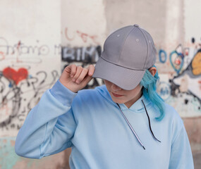 Blue haired Teenage girl in blue hoodie and baseball cap staying against graffiti wall