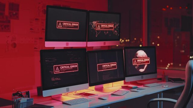 Zooming in of critical error warning message showing on multiple computer screens on desk in office at night, red light flashing