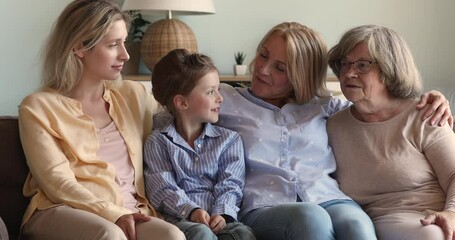 Multigenerational women talking sit on sofa at home, hugging having pleasant warm conversation. 6s girl her young mom, grandmother and great-grandmother spend time together. Trust, family bond concept