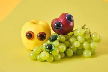 plum, apple and bunch of grapes with puppet eyes, prank concept, green background, cute fruit faces