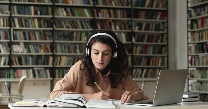 Concentrated millennial Hispanic female in headset study at library prepare essay coursework using laptop. Focused young lady student make research write up information from books and online sources