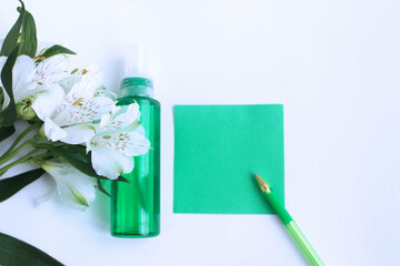 A green transparent bottle with cosmetics, white alstroemeria flowers and a sheet of green writing paper on a white background. Cosmetic products.