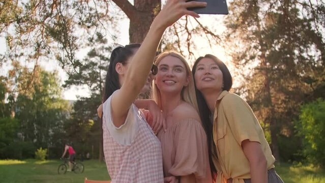 Group of three attractive young women wearing stylish casual clothes standing in park taking selfie on smartphone