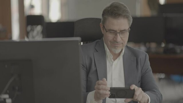 Business portrait - confident businessman sitting at desk working in office, using phone. Happy mid adult man in shirt and jacket, smiling. Video in LOG format - S-LOG3 Cine.