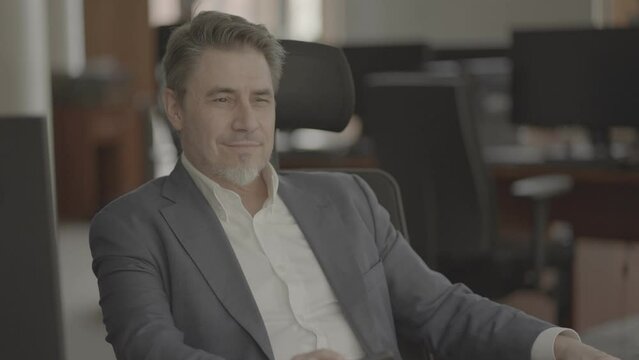 Business portrait - confident businessman sitting at desk in office. Happy mid adult man in shirt and jacket, smiling. Bearded, gray hair. Video in LOG format - S-LOG3 Cine.