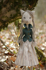 Cute blonde doll cat with green eyes dressed in a  jacket and nice dress standing next to a tree.
