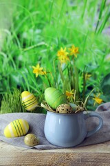 Easter eggs, a cup with Easter decorations and a flower, on a wooden table, against the background of green grass and blooming yellow daffodils, the concept of home celebrations, congratulations, post