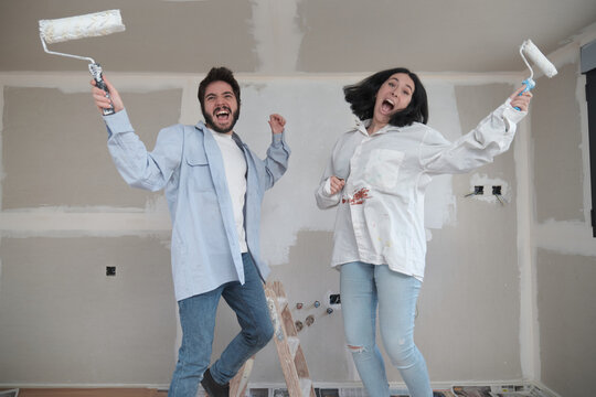 Young Happy Caucasian Couple Jumping With Paint Rollers And Laughing While Painting Walls In Their New House In Construction.