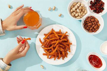 Womans hands take sweet potato fries, nuts and drinks on blue background with long shadows in...