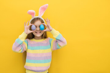 A little happy girl with long blonde hair in a colorful bright sweater with a hare's ears is holding colorful Easter eggs on a yellow background, an Easter card, an Easter banner, a place for text