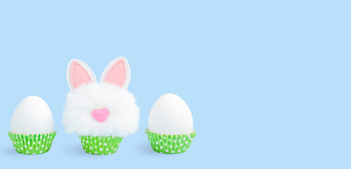 Unpainted Easter eggs in green paper cake pans and funny easter bunny toy on blue background. Banner with copy space