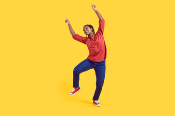 Fototapeta na wymiar Happy cheerful young black girl having fun in the studio. Funny African American woman in comfortable casual clothes dancing against a vivid, vibrant yellow colour background in a modern photo studio