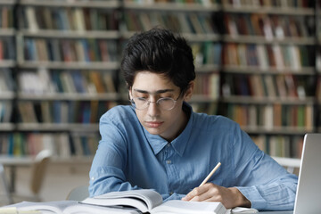 Focused busy young male Jewish student in eyeglasses reading educational textbook, improving knowledge writing notes, preparing for exam or doing homework alone in library, learning concept.